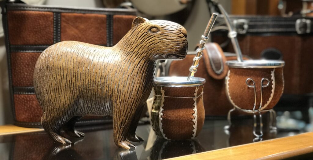 A carved wooden capybara figure next to two leather-wrapped traditional Yerba Mate gourds with stainless steel bombilla straws on a glass display shelf.