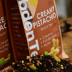 A close-up shot of dry, loose-leaf Creamy Pistachio black tea blend from Hebden Tea, featuring dark black tea leaves mixed with real pistachio pieces, sweet dates, yogurt granules, and bright pink cornflower petals.