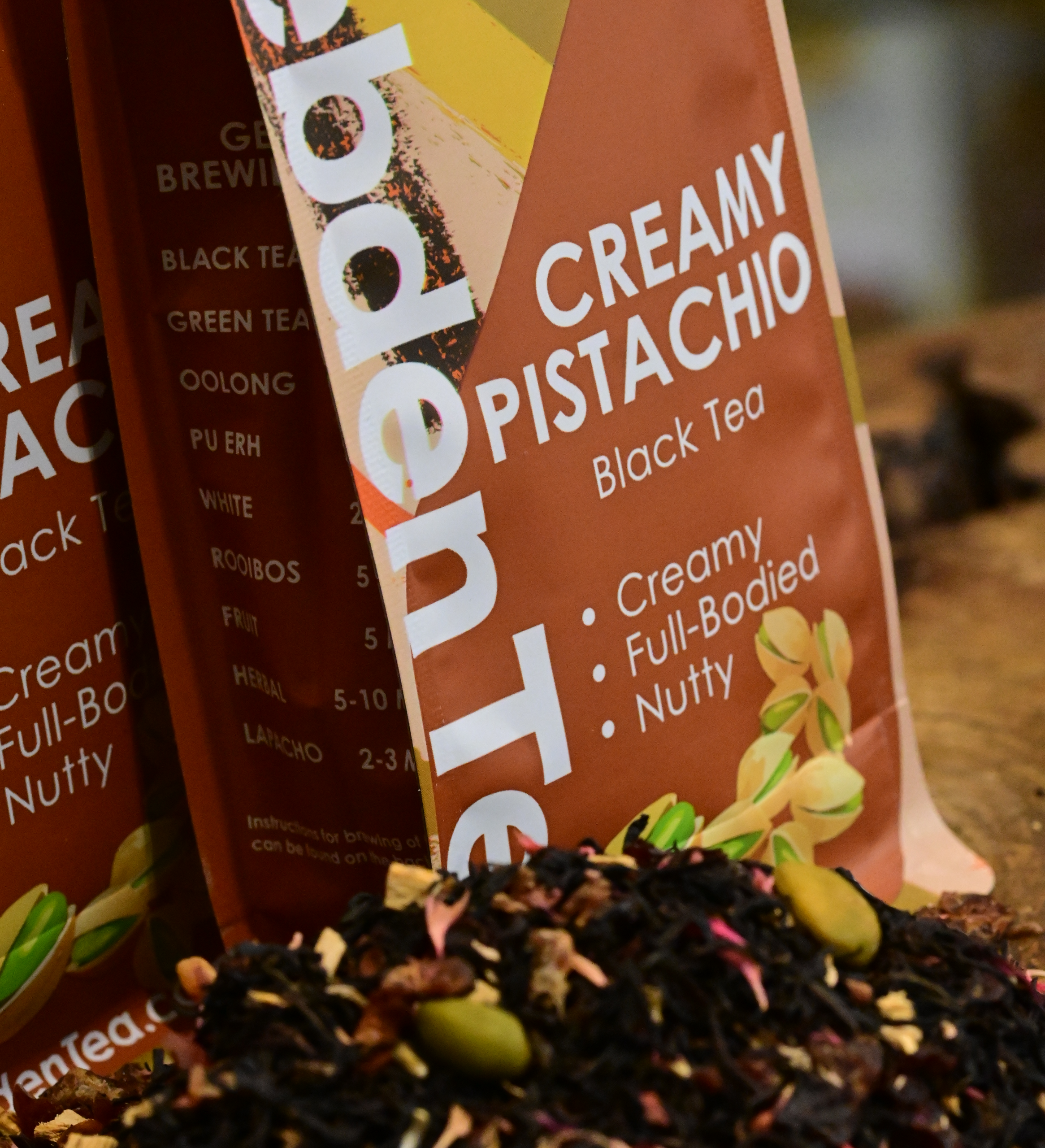 A close-up shot of dry, loose-leaf Creamy Pistachio black tea blend from Hebden Tea, featuring dark black tea leaves mixed with real pistachio pieces, sweet dates, yogurt granules, and bright pink cornflower petals.